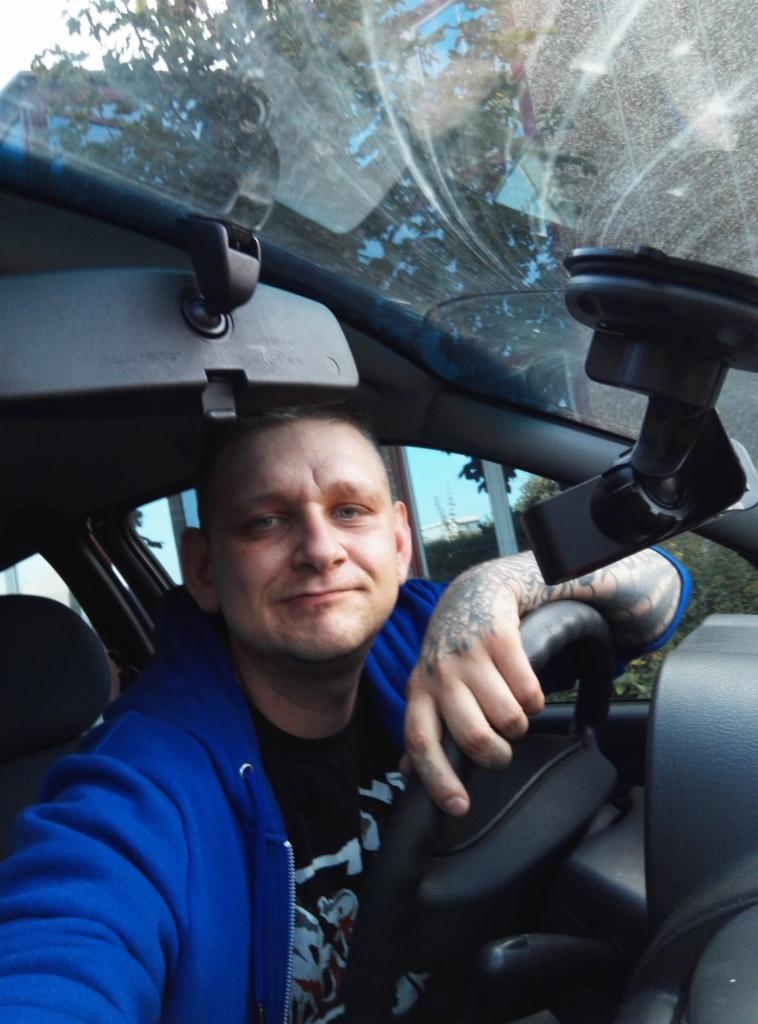 Mężczyzna w niebieskiej bluzie z kapturem, z tatuażem na ręce, siedzi w samochodzie; widoczne lusterko wsteczne i uchwyt na telefon.