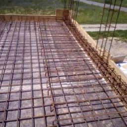 Zbrojenie płyty fundamentowej z wystającymi prętami pionowymi, widoczne szalunki drewniane i trawa w tle.