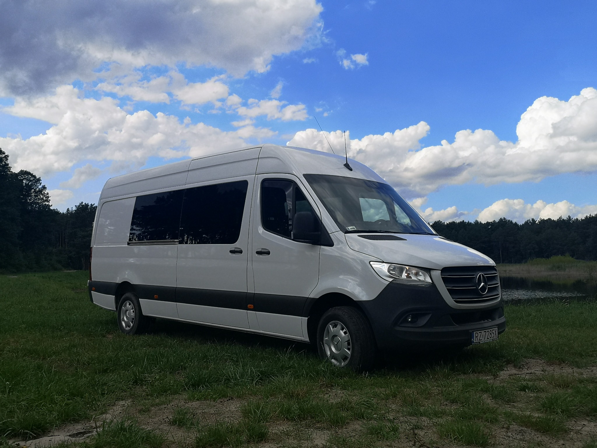 Biały bus Mercedes-Benz Sprinter z przyciemnianymi szybami na tle błękitnego nieba z obłokami, zaparkowany na trawiastym terenie obok zbiornika wodnego.