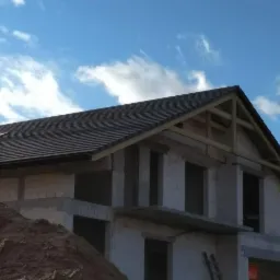 Surowy stan dwupiętrowego domu z ciemną dachówką, widoczne drewniane krokwie i komin, na pierwszym planie hałda piasku