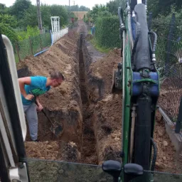 Pracownik z łopatą w wykopie obok minikoparki, przygotowanie pod przyłącze wodno-kanalizacyjne w terenie zabudowanym.