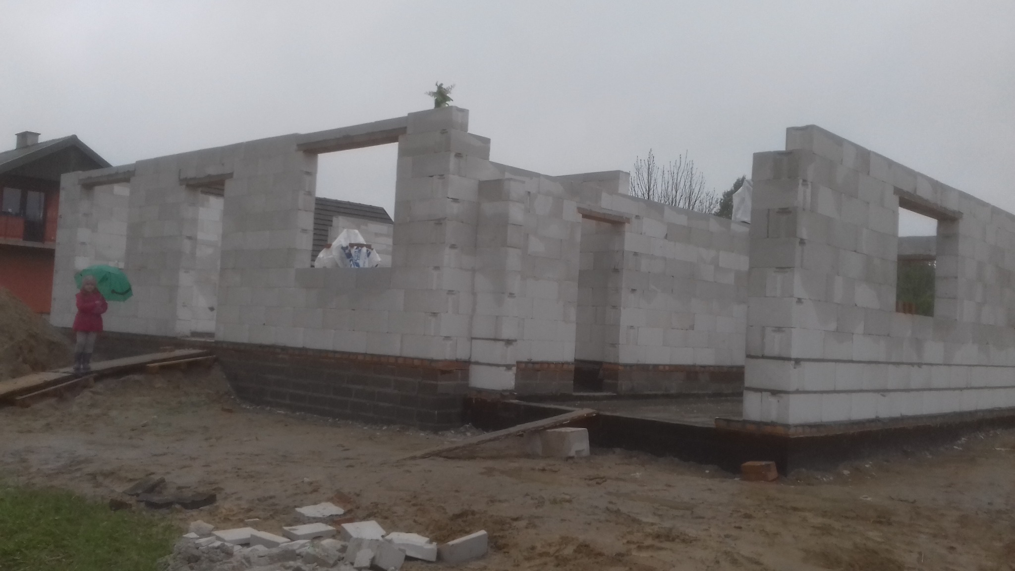 Wznoszenie ścian parterowego domu z betonu komórkowego na fundamencie z cegły, w tle osoba z zielonym parasolem i budynek.