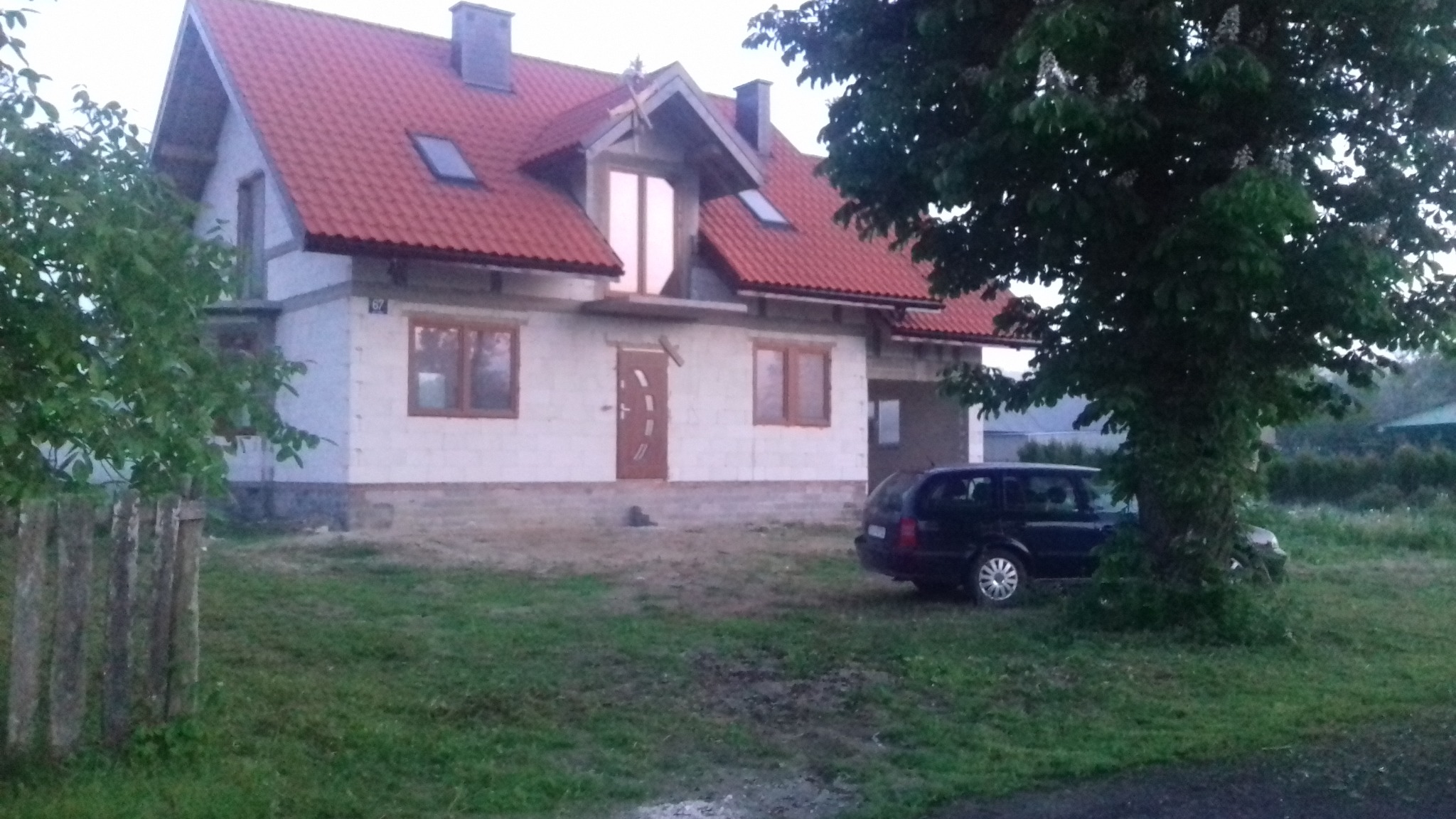 Surowy stan domu z czerwonym dachem, widoczne okna, drzwi i fundamenty, obok zaparkowany ciemny samochód, widok z zewnątrz, w tle drzewa.