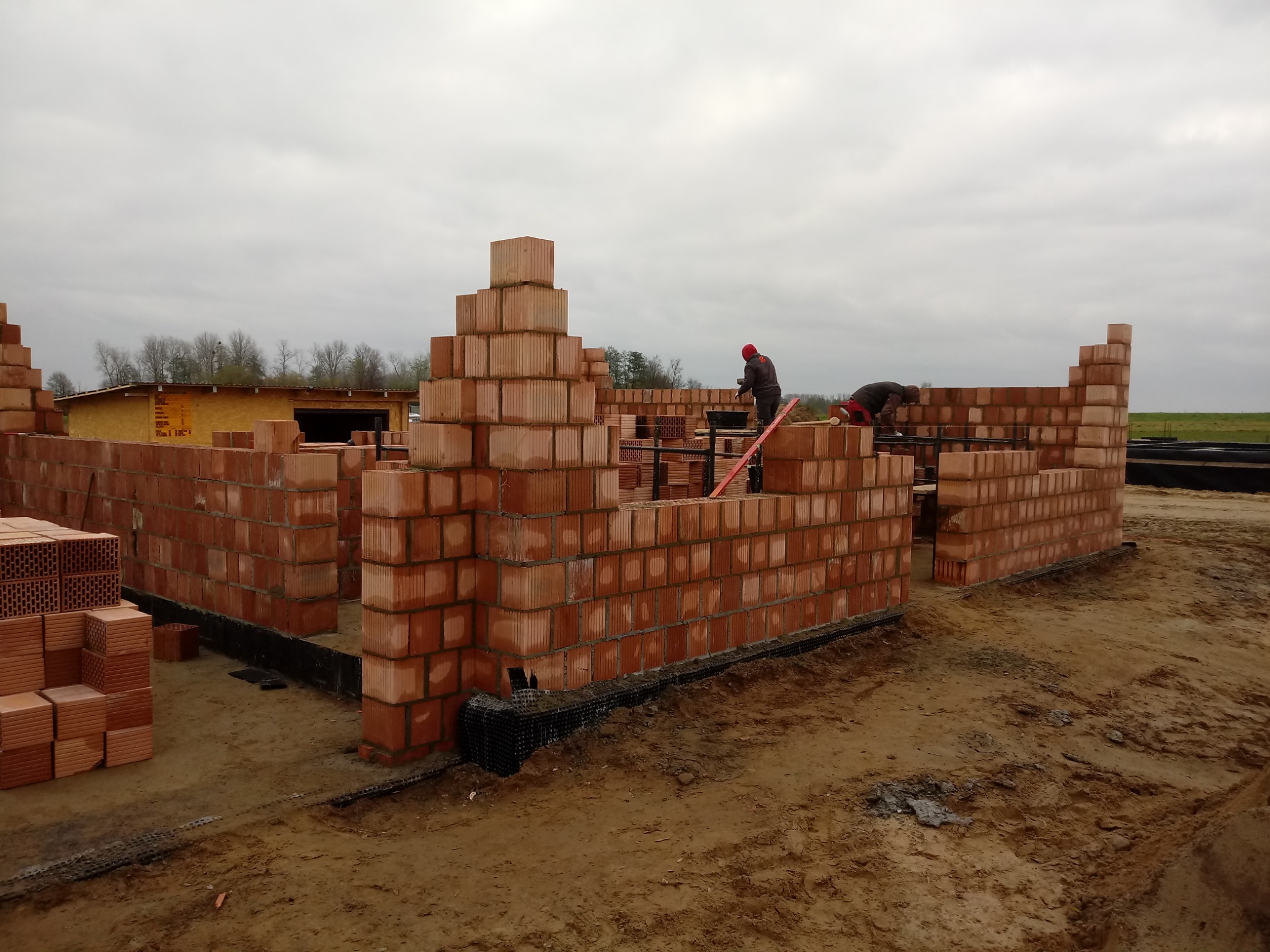 Wznoszenie ścian parterowego domu z czerwonej ceramiki poryzowanej, widoczni dwaj murarze w trakcie pracy, w tle szary krajobraz i budynek gospodarczy.