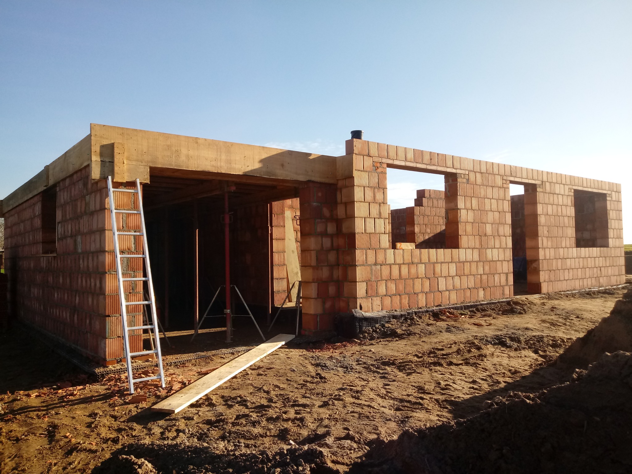 Wznoszenie ścian parterowego budynku z czerwonej cegły ceramicznej; widoczna aluminiowa drabina oparta o ścianę, drewniane nadproża okienne, szalunki stropowe i fragment niezagospodarowanego terenu.