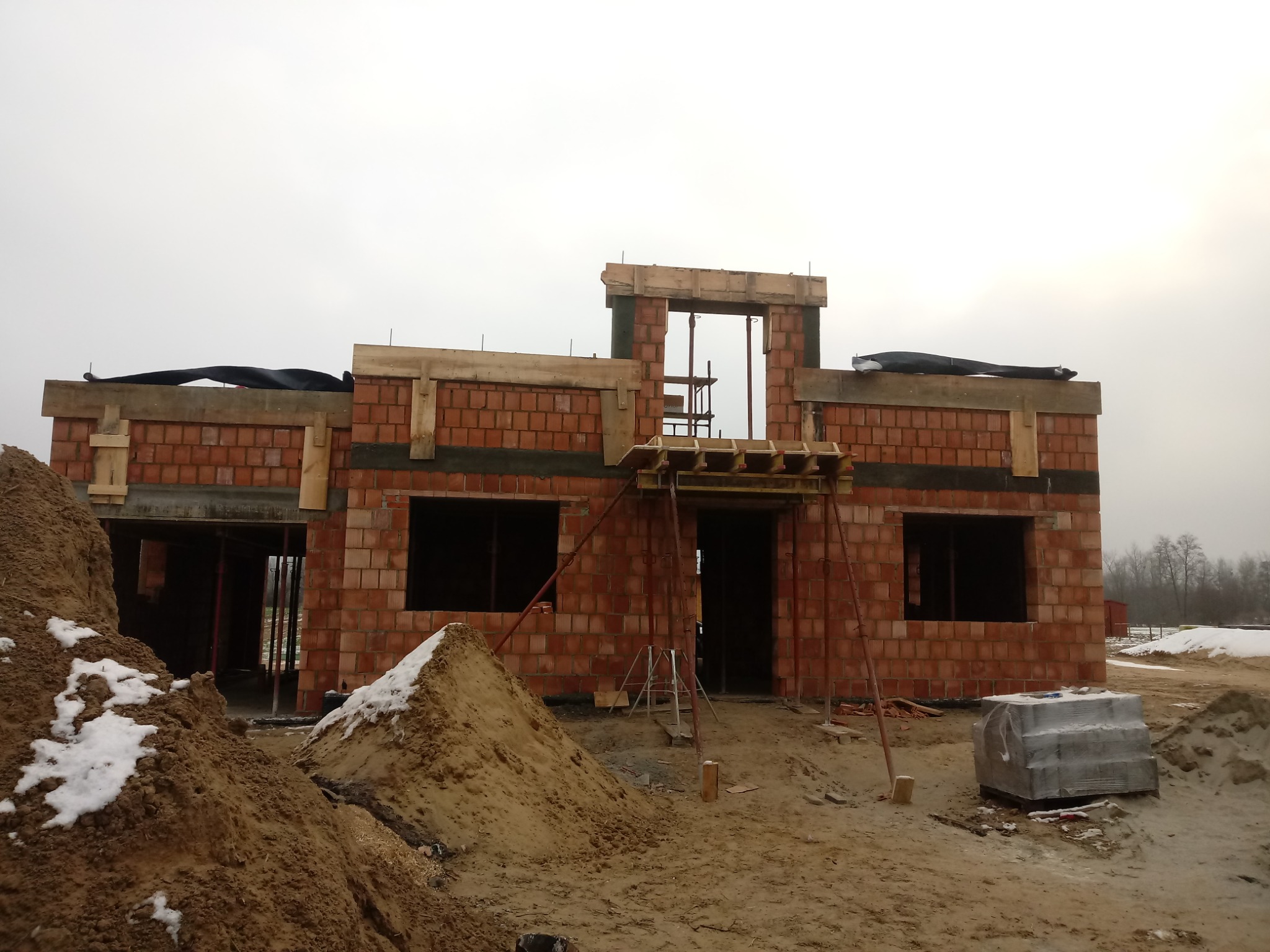 Surowy stan domu z czerwonej cegły w Cieszanowie, widoczne drewniane szalunki i podtrzymujące stemple, na pierwszym planie hałda piasku z resztkami śniegu.