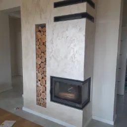 Nowoczesny kominek z obudową w odcieniach beżu, z wnęką na drewno wypełnioną pociętymi kłodami, czarny wkład kominkowy i dekoracyjne czarne elementy w górnej części obudowy.