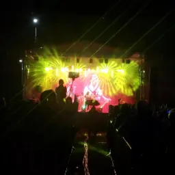 Koncert na scenie z intensywnym oświetleniem i laserami, widoczna sylwetka muzyka i publiczność w ciemności.