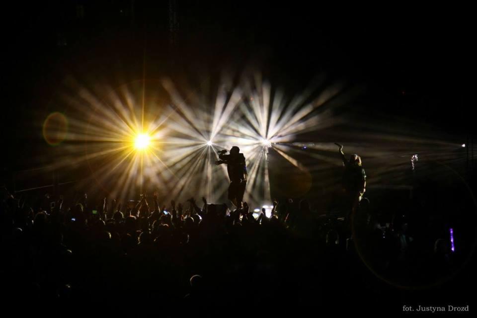 Koncert z sylwetkami artystów na scenie oświetlonej reflektorami, tłum publiczności z podniesionymi rękami w ciemności.