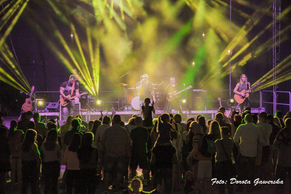 Zespół muzyczny na scenie podczas koncertu plenerowego, oświetlony reflektorami i otoczony tłumem fanów.