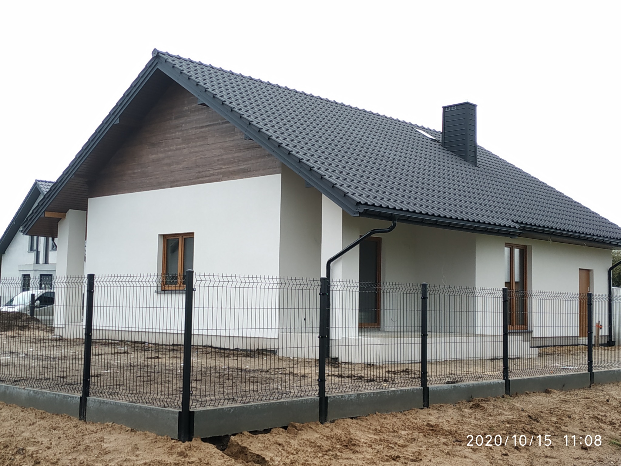 Nowoczesny dom jednorodzinny z białą elewacją, grafitową dachówką i drewnianym panelem na szczycie, otoczony metalowym ogrodzeniem panelowym na betonowej podmurówce, teren wokół w trakcie...