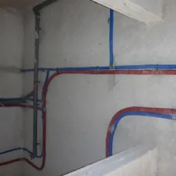 Surowe ściany z fragmentami instalacji hydraulicznej w kolorze niebieskim i czerwonym, rury połączone metalowymi zaciskami, widoczny szary pion kanalizacyjny.