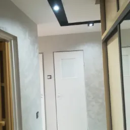 Korytarz po odświeżeniu, ściany pokryte dekoracyjnym tynkiem w odcieniach szarości, białe drzwi z przeszkleniem, czarny pas z oświetleniem punktowym na suficie, widoczna część szafy z przesuwnymi...