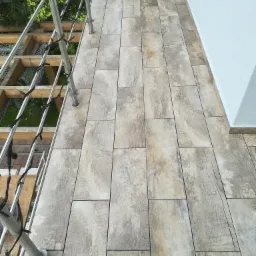 Ułożone płytki imitujące drewno na balkonie, widoczna metalowa balustrada i fragment elewacji budynku.