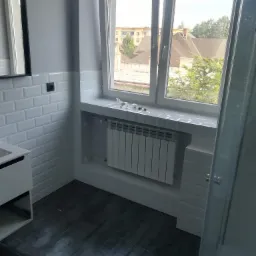 Wykończona łazienka z białymi płytkami typu metro, ciemną podłogą, grzejnikiem pod oknem i widokiem na budynki oraz drzewa za oknem.