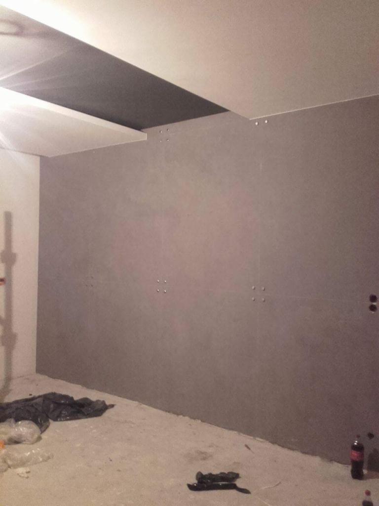 Pokój w trakcie remontu: szara ściana z widocznymi łączeniami płyt gipsowo-kartonowych, sufit z fragmentem pomalowanym na ciemny kolor, na podłodze resztki materiałów budowlanych i butelka po napoju.