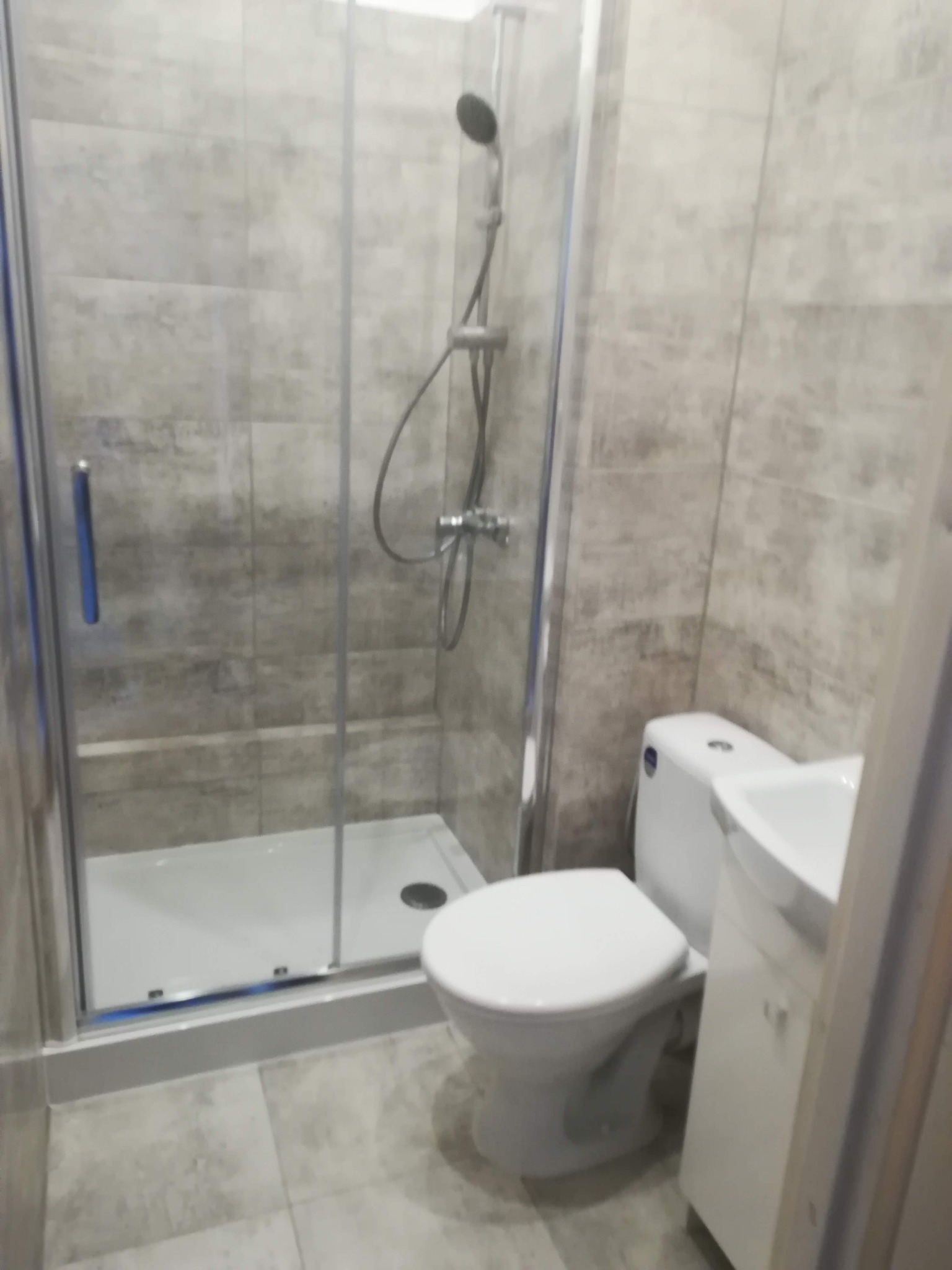 Nowoczesna łazienka z kabiną prysznicową z przesuwnymi drzwiami, umywalką z szafką i toaletą, wykończona płytkami w odcieniach szarości.