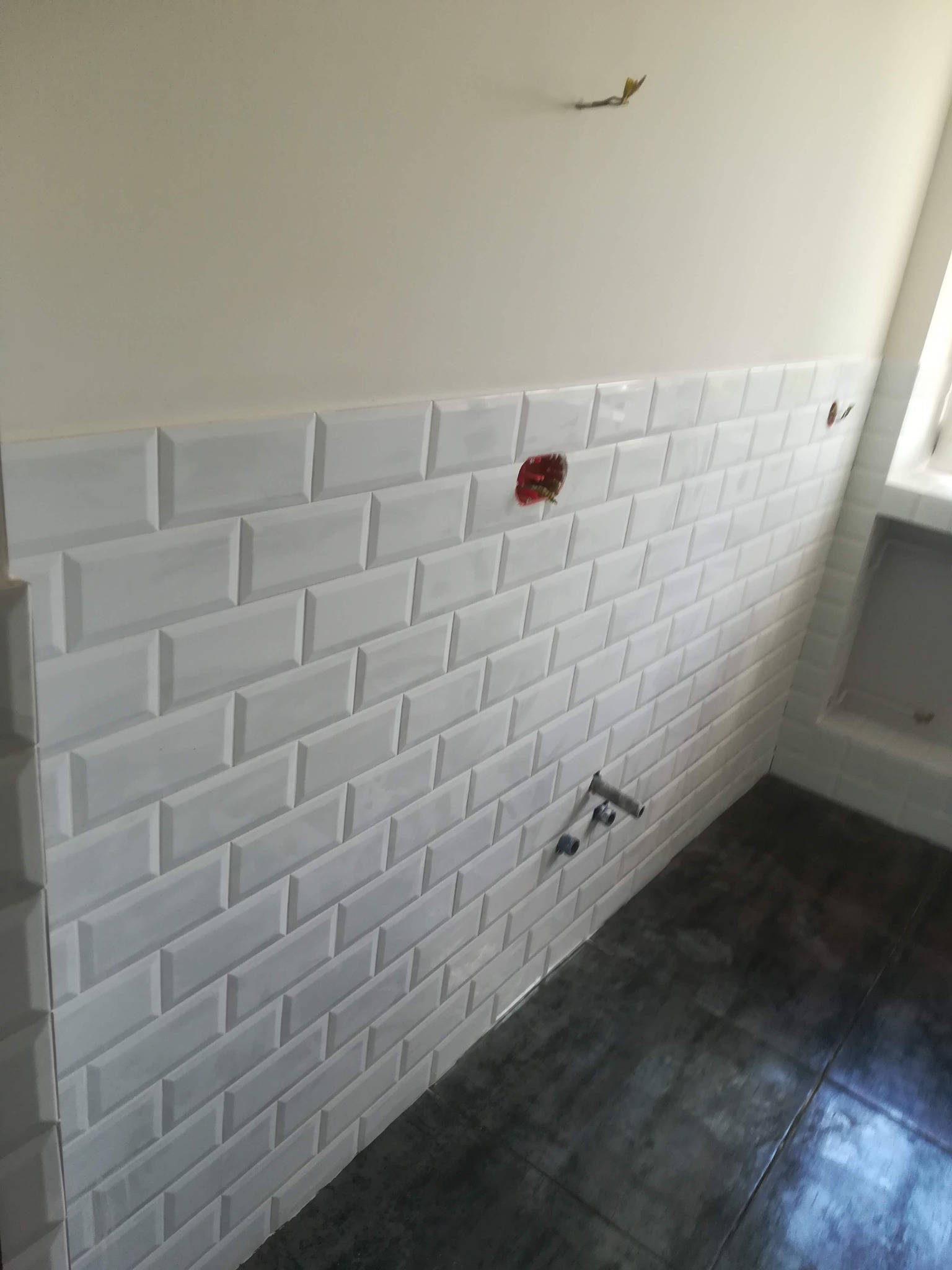 Ściana w łazience wyłożona białymi, błyszczącymi płytkami typu metro, z widocznymi przyłączami wodnymi i puszką elektryczną przed montażem osprzętu, szara podłoga z fugami.