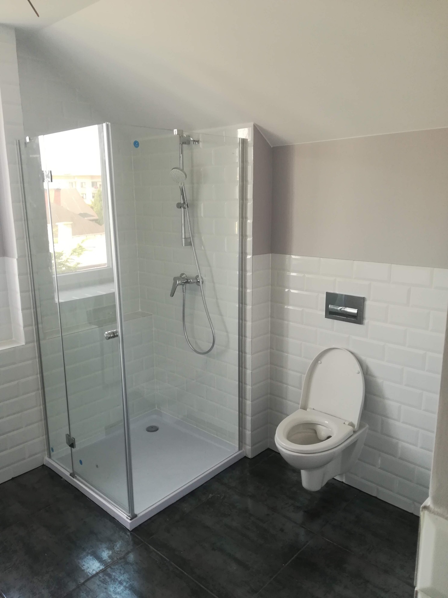 Nowoczesna łazienka na poddaszu z kabiną prysznicową ze szkła i białymi płytkami typu metro, wisząca toaleta z chromowanym przyciskiem spłukiwania, ciemna podłoga.