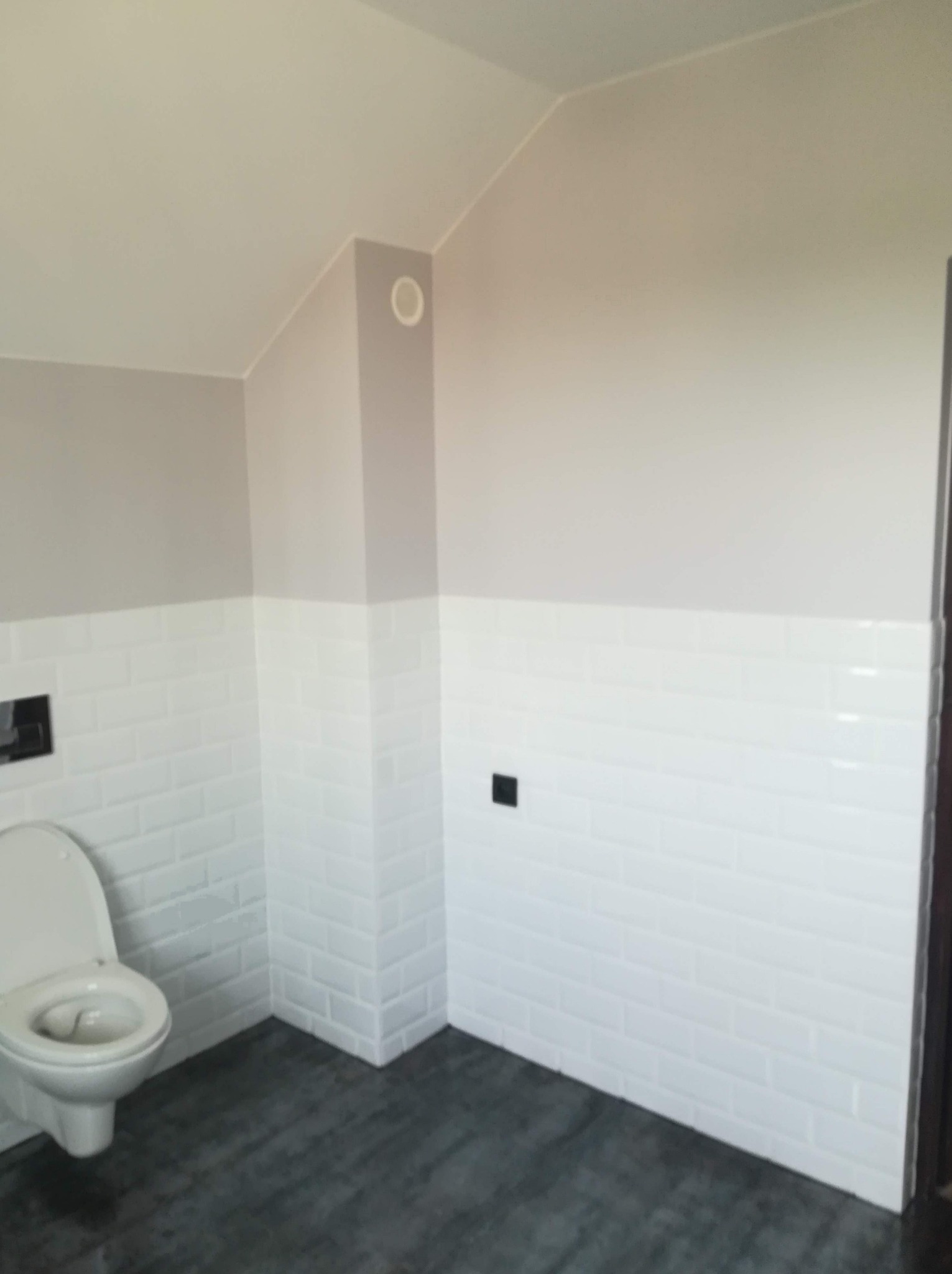 Wykończona łazienka w odcieniach szarości i bieli, z toaletą, białymi płytkami typu metro i ciemną podłogą imitującą drewno, widoczny fragment wentylacji.