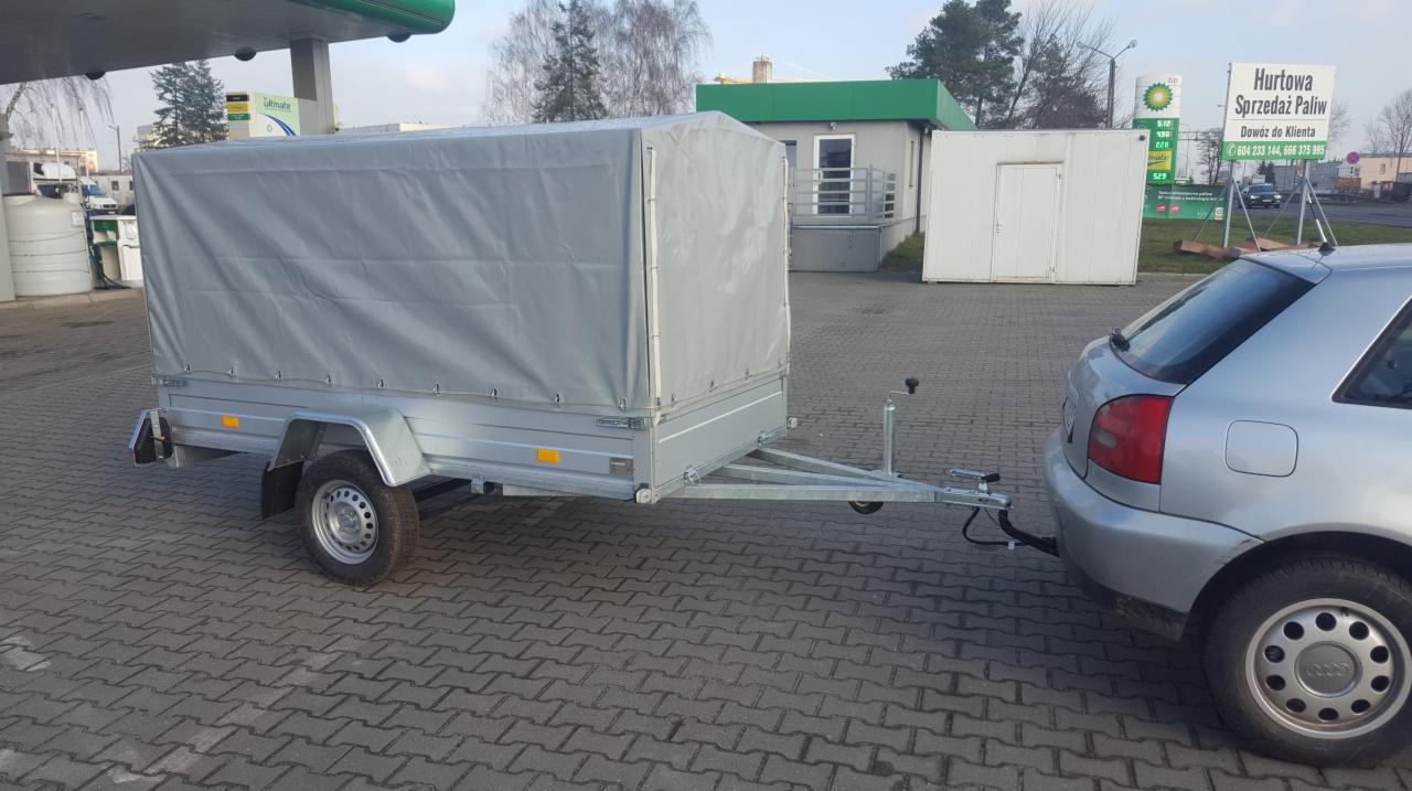 Szara przyczepa z plandeką podpięta do srebrnego samochodu osobowego na tle stacji benzynowej i budynku biurowego.