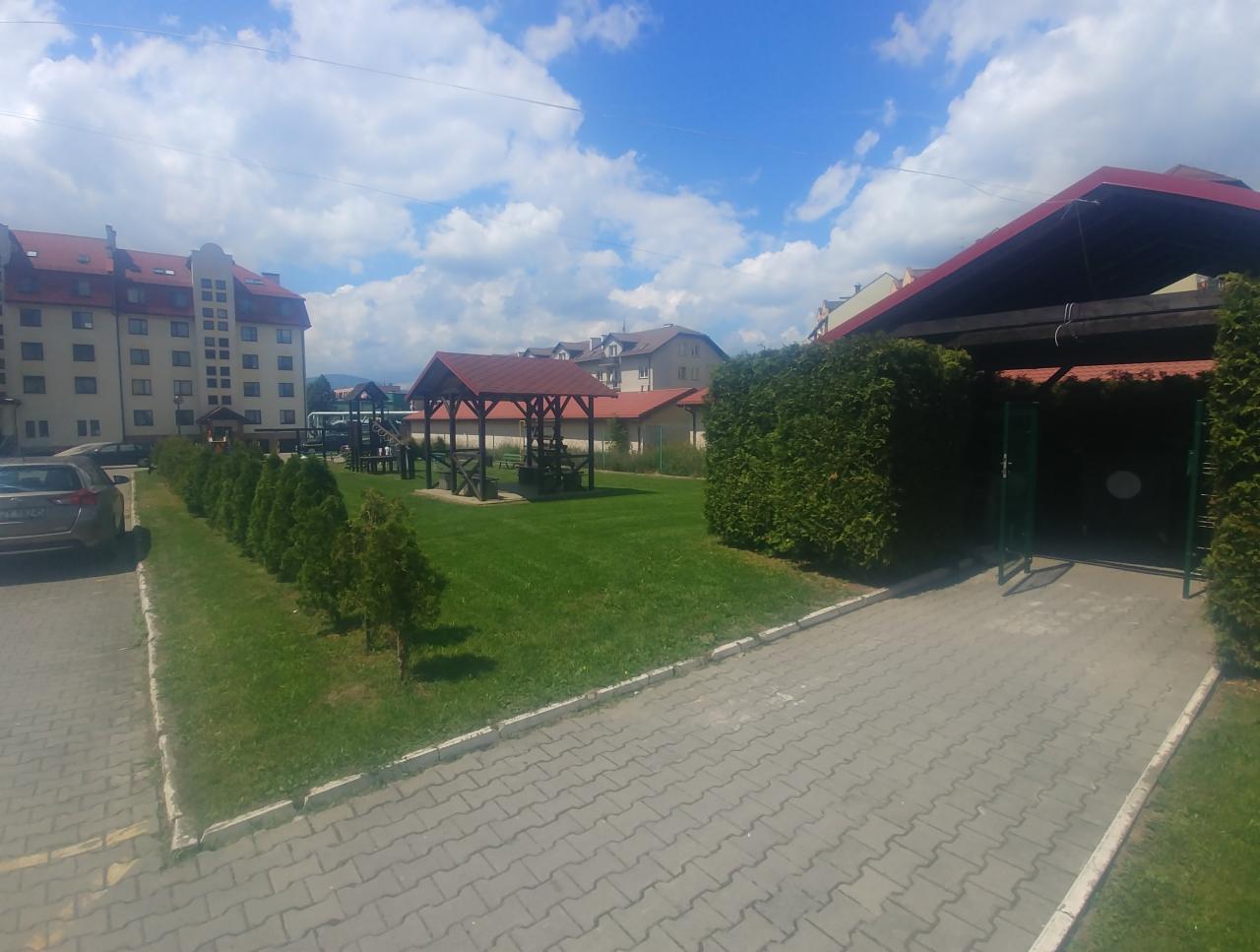 Uporządkowany teren zielony z rzędem młodych drzewek iglastych, placem zabaw pod zadaszeniem i budynkiem mieszkalnym w tle, widziany z perspektywy chodnika wyłożonego kostką brukową.