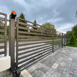 Nowoczesna, przesuwna brama wjazdowa z automatyką, z szarymi, poziomymi panelami, pomarańczową lampą ostrzegawczą i napędem liniowym, na tle brukowanej nawierzchni i zieleni.