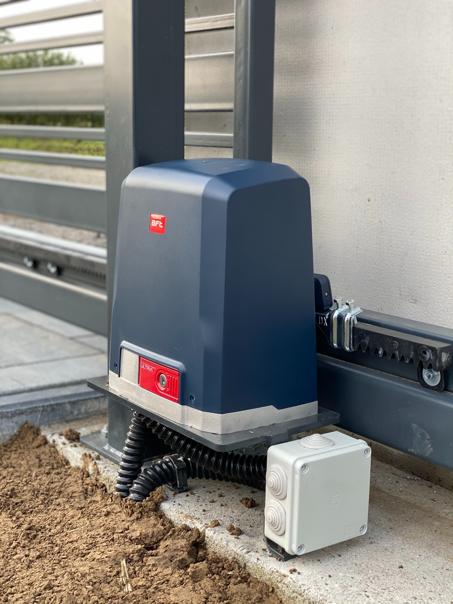 Zamontowany napęd BFT do bramy przesuwnej, widoczna skrzynka sterownicza i przewody w peszlach, świeżo zasypana ziemia wokół.