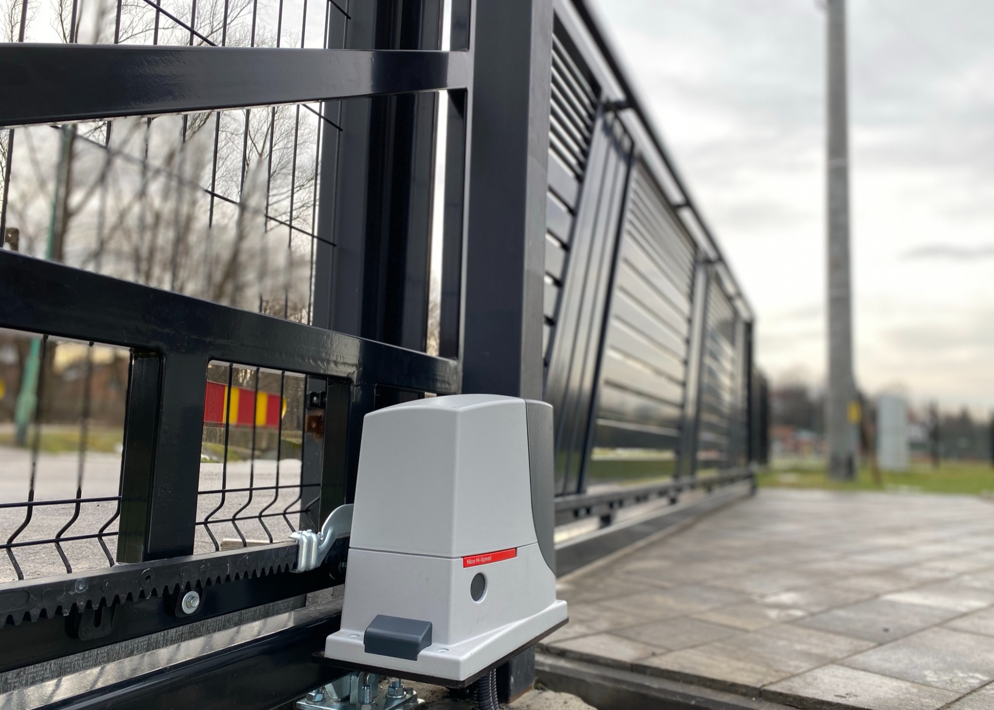 Zamknięta, przesuwna brama automatyczna z widocznym napędem Nice Hi-Speed zamontowanym na betonowej kostce brukowej, z elementami metalowymi i siatką w tle.