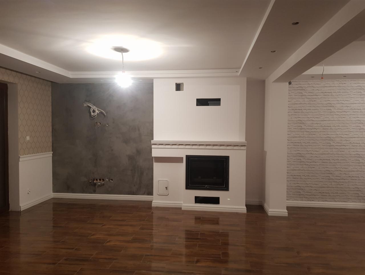 Wnętrze w trakcie wykończenia: ściana z efektem betonu z widocznymi przewodami, kominek obudowany białą ramą, fragment ściany wyłożonej białą cegłą, parkiet w odcieniach brązu.