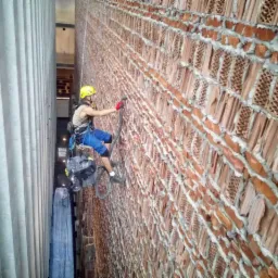 Pracownik w kasku, zabezpieczony linami, czyści ceglaną elewację budynku za pomocą urządzenia pod ciśnieniem. Widoczna kolumna budynku po lewej stronie.