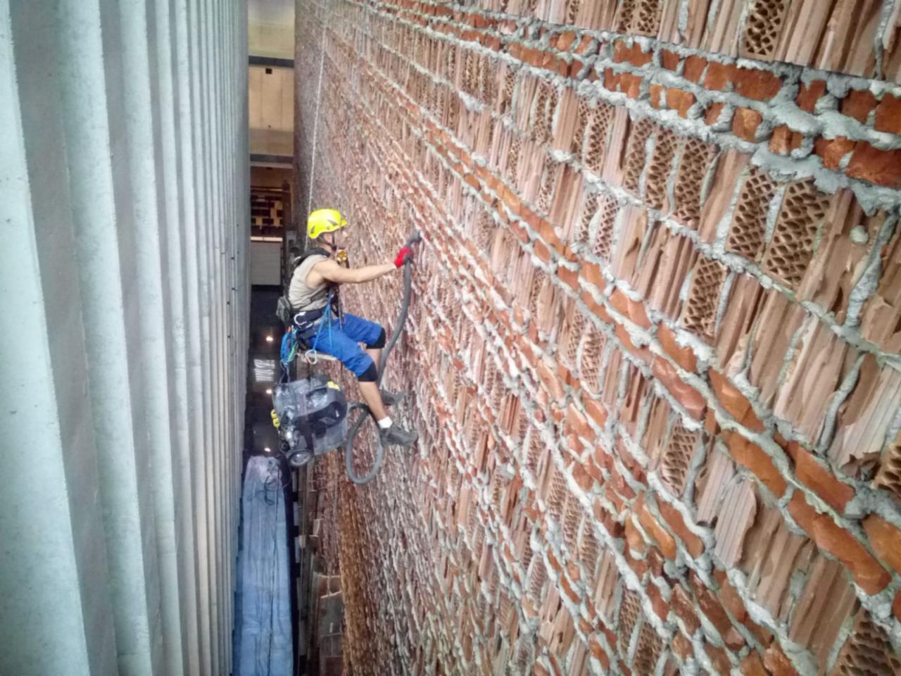 Pracownik w kasku, zabezpieczony linami, czyści ceglaną elewację budynku za pomocą urządzenia pod ciśnieniem. Widoczna kolumna budynku po lewej stronie.