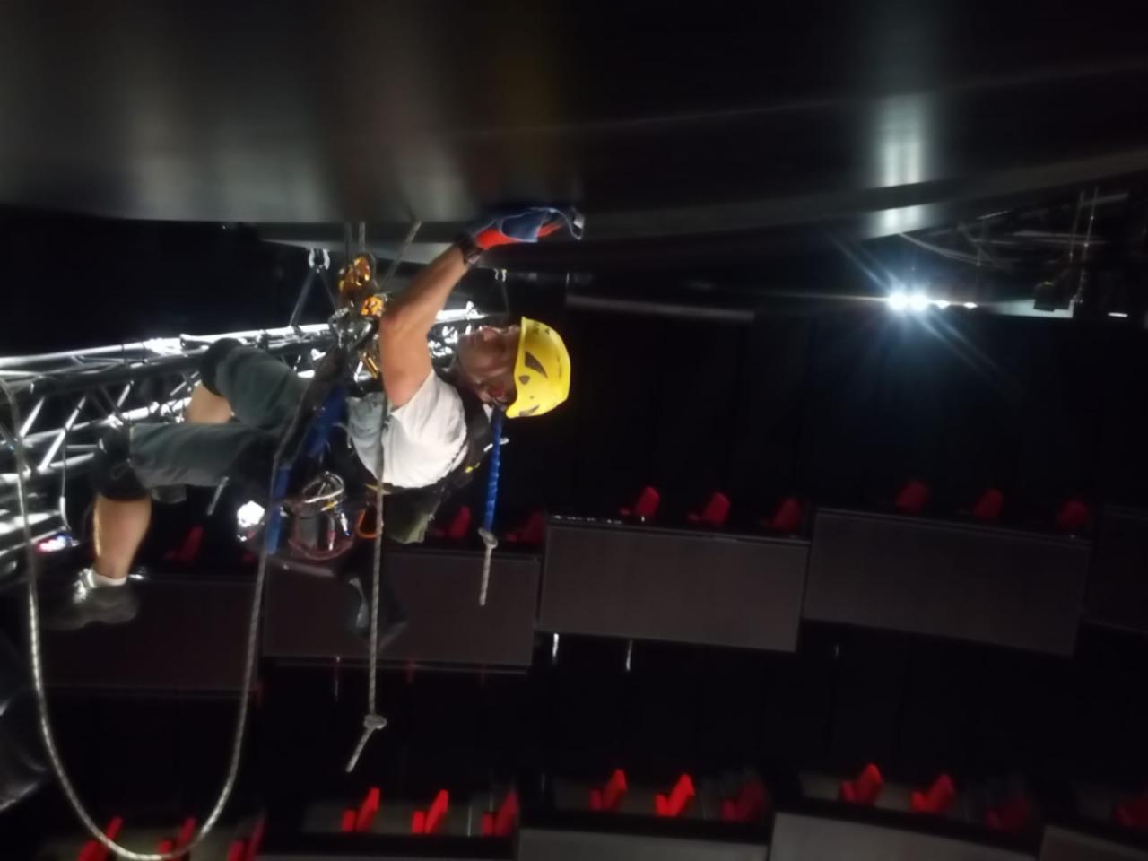 Pracownik w uprzęży, z żółtym kasku, czyści sufit hali widowiskowej z reflektorami, używając szmatki i lin.