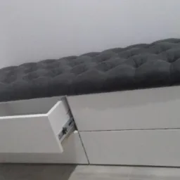 Tapicerowana ławka z szufladami na wymiar, białe fronty, ciemnoszara tapicerka z pikowaniem, jedna szuflada wysunięta.