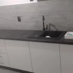 Nowoczesna kuchnia z białymi szafkami, ciemnym blatem i dwoma zlewozmywakami: czarnym i białym, pod szarymi kaflami.