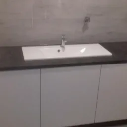 Biała, prostokątna umywalka z chromowaną baterią na blacie z ciemnego konglomeratu, osadzona na szafce z białymi frontami, widoczna na tle szarych płytek ściennych.