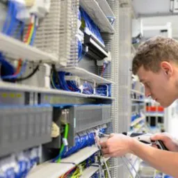 Elektryk w niebieskiej koszulce podłącza przewody w rozdzielnicy; widoczne liczne kable, listwy zaciskowe i elementy sterujące.