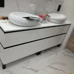 Biała szafka łazienkowa z dwiema okrągłymi umywalkami nablatowymi, widoczne narzędzia i akcesoria montażowe, marmurowe płytki na ścianie i podłodze, etap montażu.