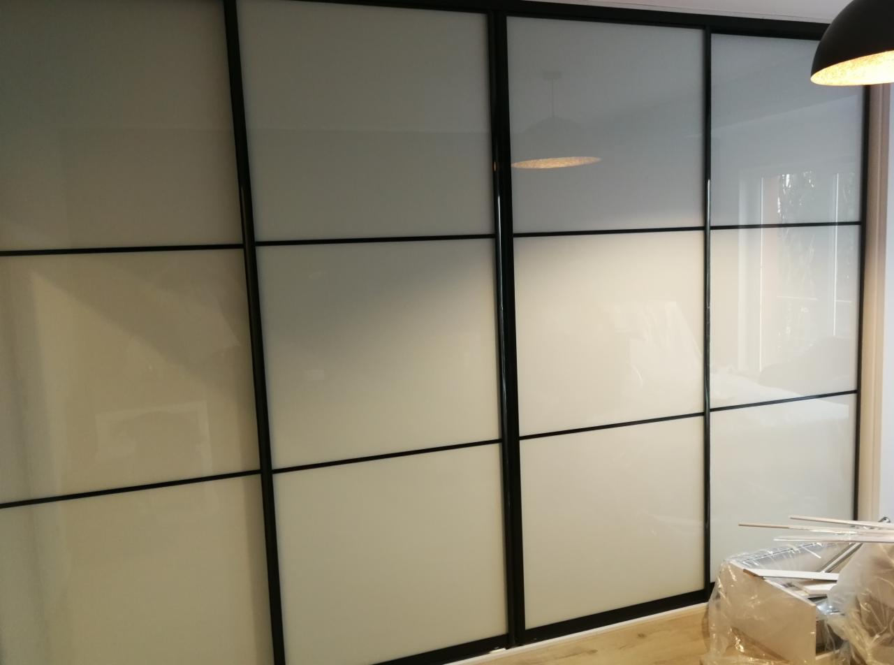 Czterodrzwiowa szafa przesuwna z białymi, matowymi frontami i czarnymi ramami, widoczna lampa sufitowa i częściowo rozpakowane elementy montażowe na podłodze.