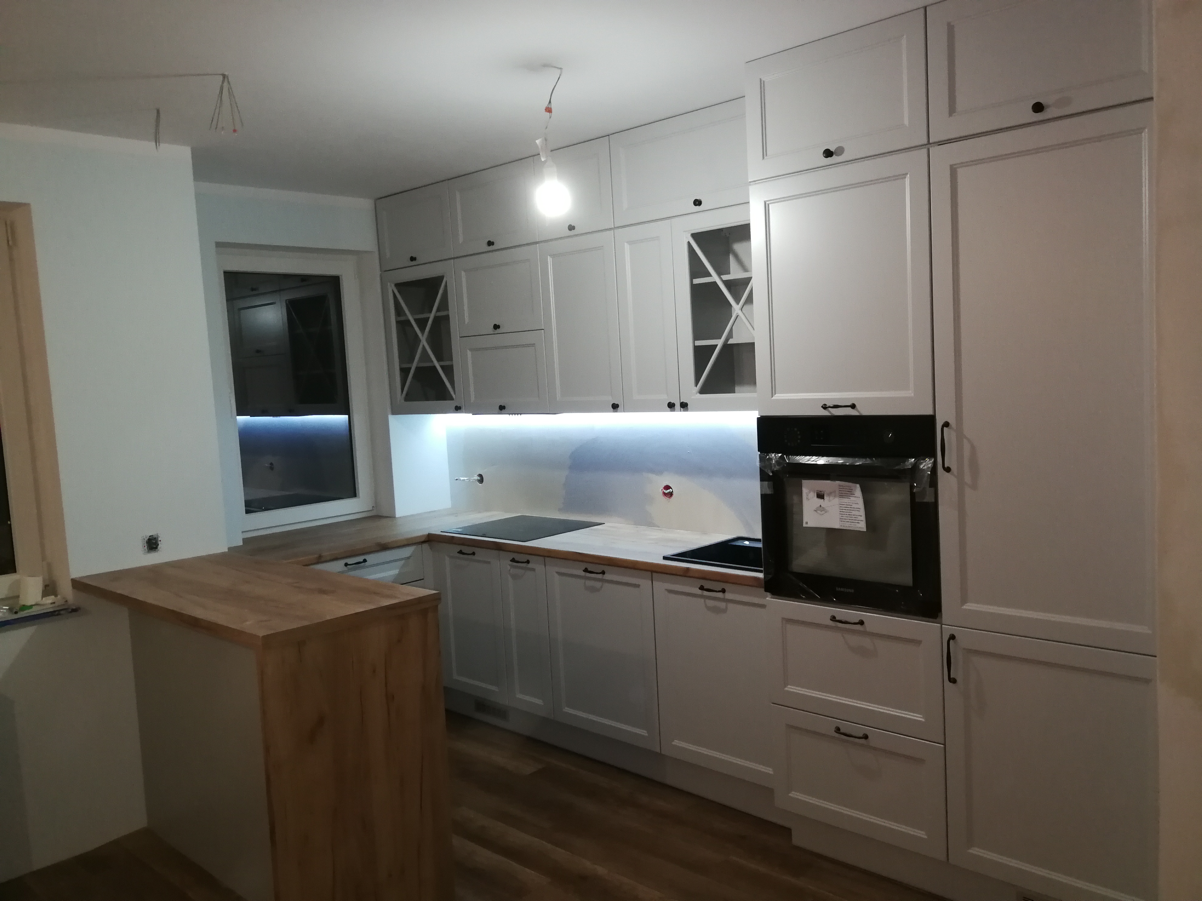 Jasnoszara zabudowa kuchenna z drewnianym blatem i barkiem, widoczne podświetlenie LED pod szafkami wiszącymi, piekarnik w folii ochronnej, surowa żarówka zwisająca z sufitu.