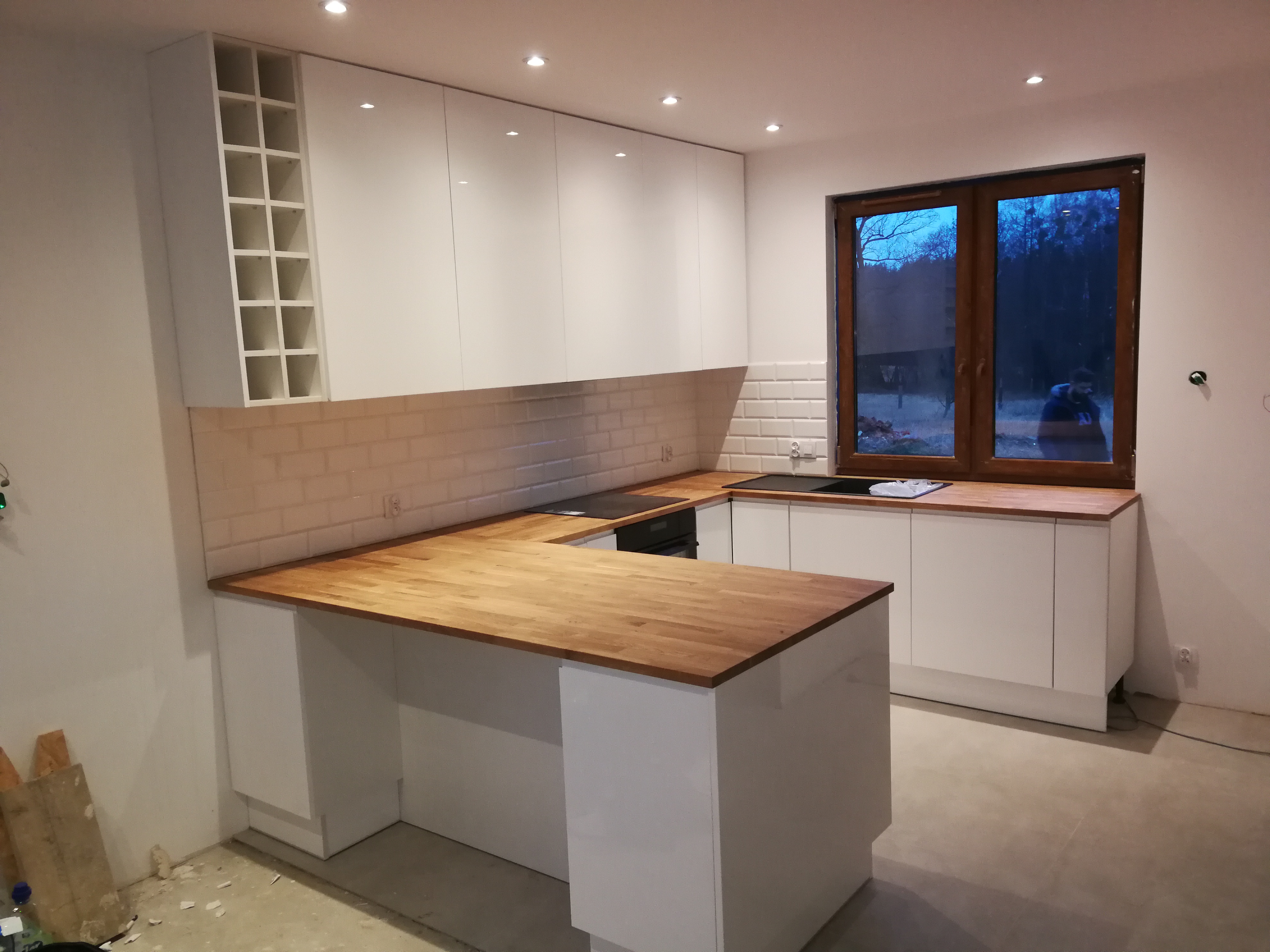 Nowoczesna biała kuchnia z drewnianym blatem, widok na okno z widocznym drzewem, niedokończona podłoga i zabudowa z półkami na wino.