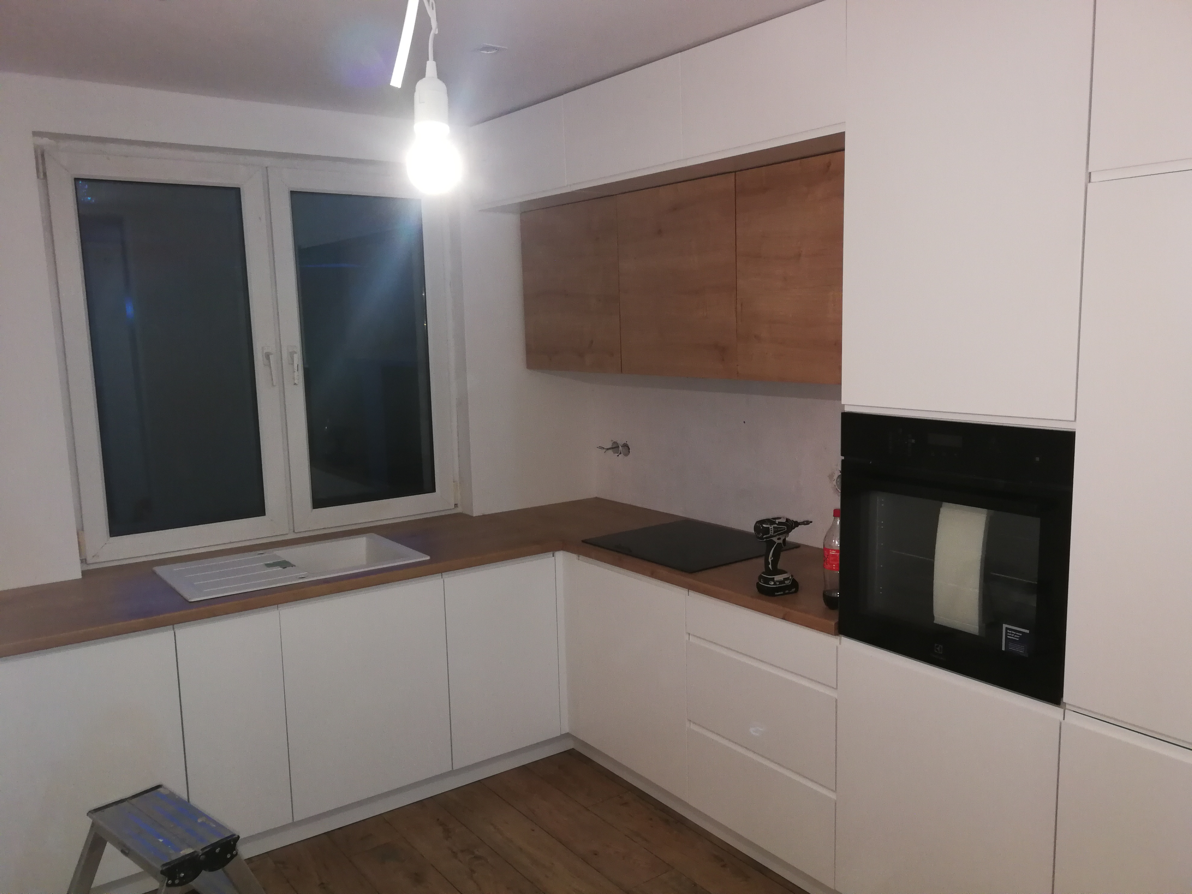 Biała kuchnia z drewnianym blatem i górnymi szafkami w kolorze drewna; w tle okno, na blacie wiertarka i butelka, na podłodze stołek.