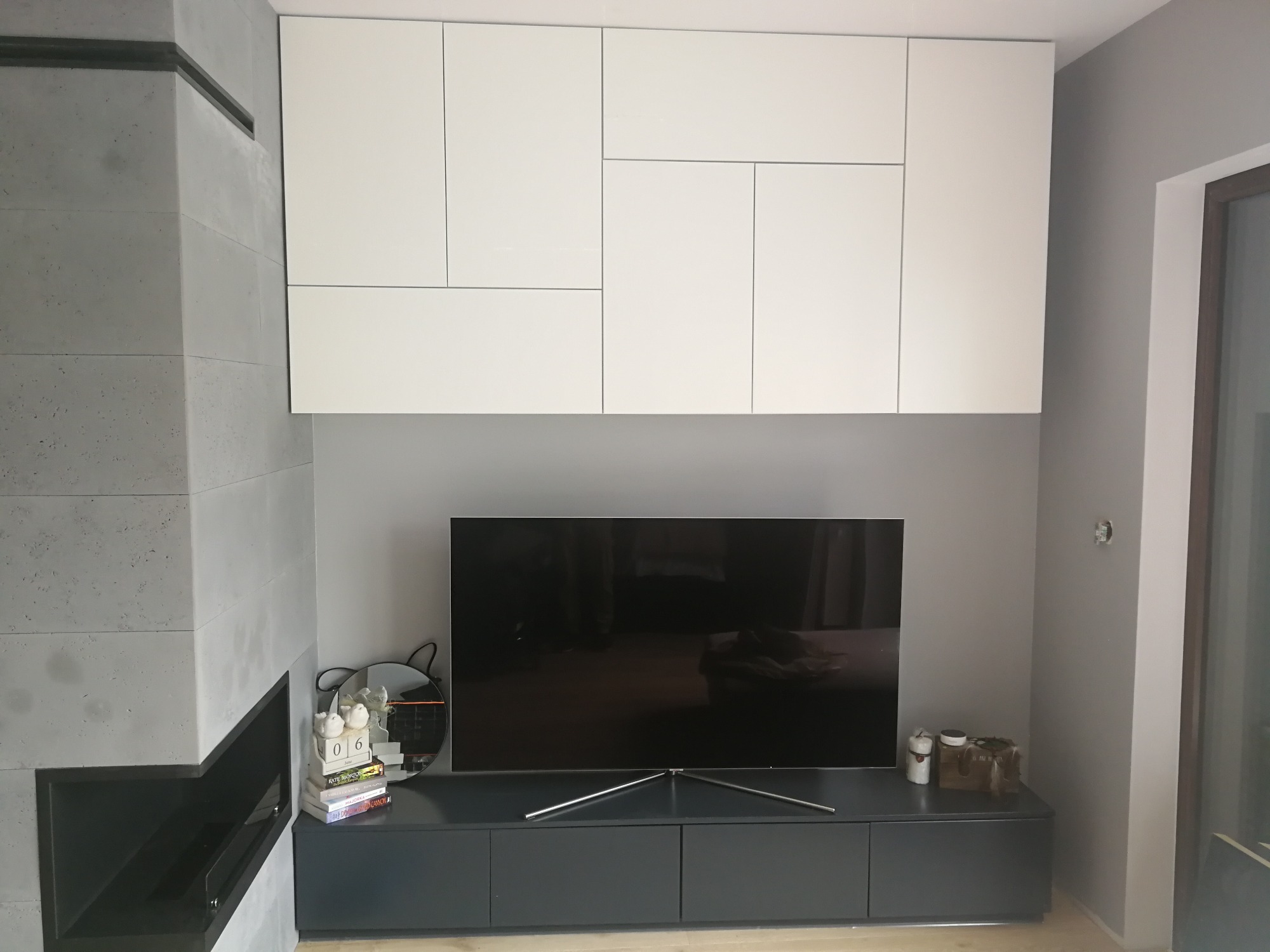 Nowoczesny salon z szarą ścianą, zabudową kominkową z elementami betonu architektonicznego, szafką RTV w kolorze grafitowym, telewizorem oraz białymi szafkami wiszącymi o asymetrycznym układzie...