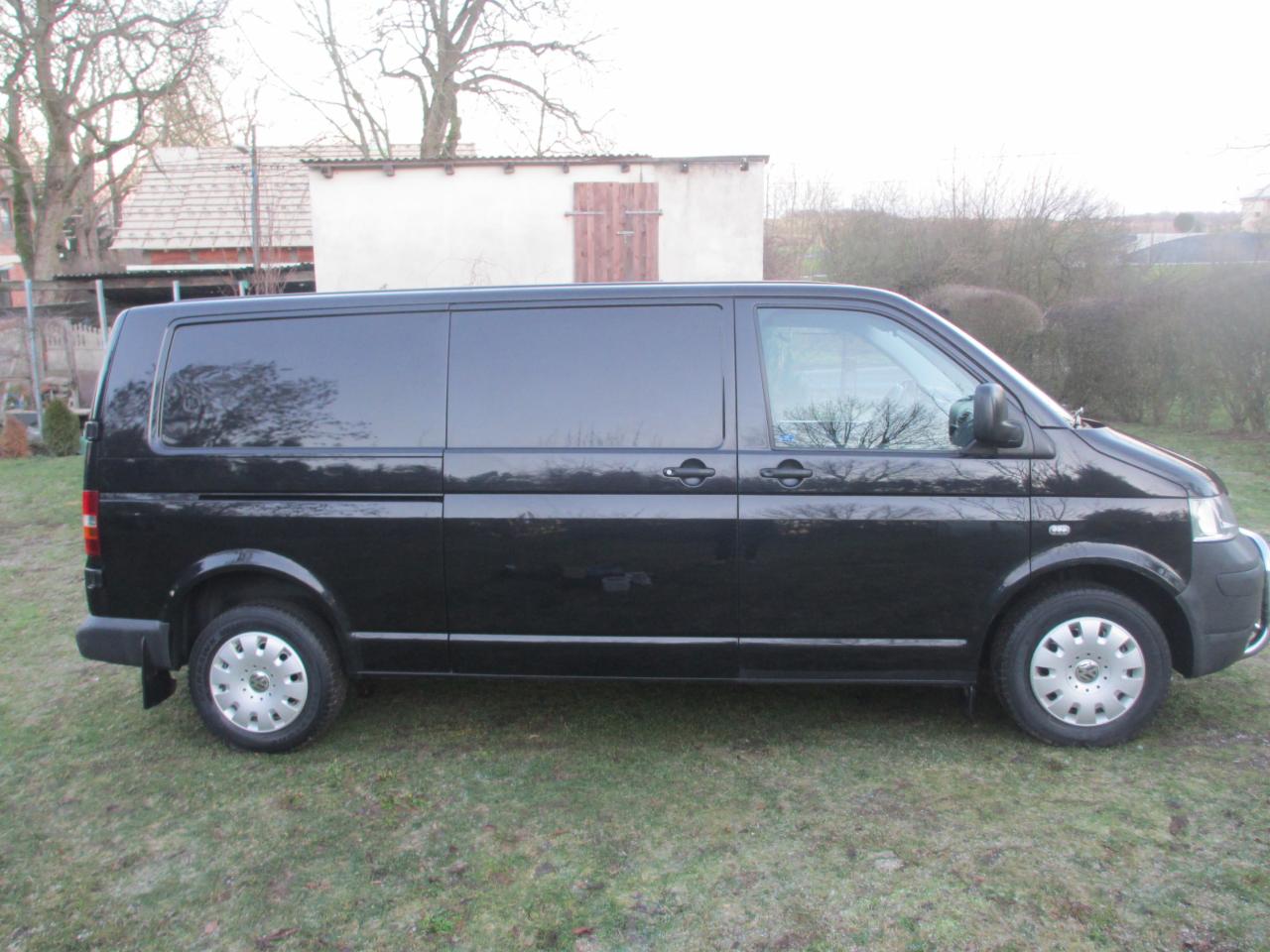 Czarny bus Volkswagen Transporter T5 z przyciemnionymi szybami, zaparkowany na trawie przed budynkiem gospodarczym z drewnianymi drzwiami.