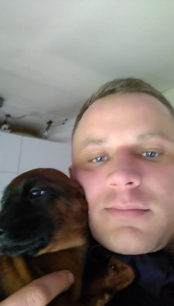 Mężczyzna z niebieskimi oczami obejmuje szczeniaka z brązową sierścią na tle białego sufitu z oświetleniem.