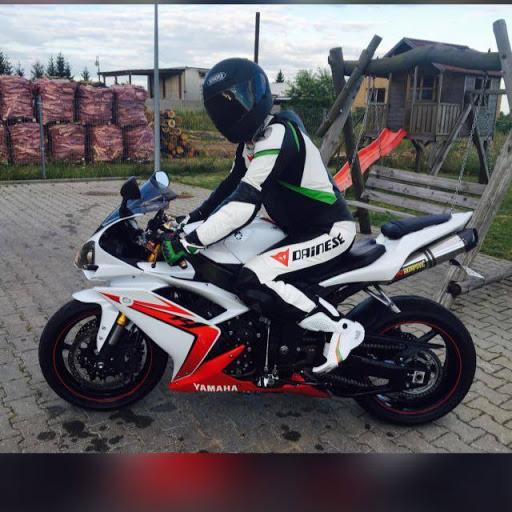 Motocyklista w kombinezonie Dainese siedzący na białym motocyklu Yamaha R1 na tle placu zabaw i sterty drewna.