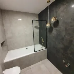 Nowoczesna łazienka z wanną obudowaną płytkami w odcieniach szarości, szklanym parawanem nawannowym z czarną ramą oraz złotą armaturą, uzupełniona o wiszące lampy w kształcie kul i toaletę w rogu.