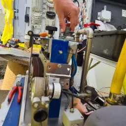 Pracownik z tatuażem na ramieniu reguluje maszynę do obróbki kabli telekomunikacyjnych w przenośnym stanowisku pracy, widoczne narzędzia i elementy instalacji.