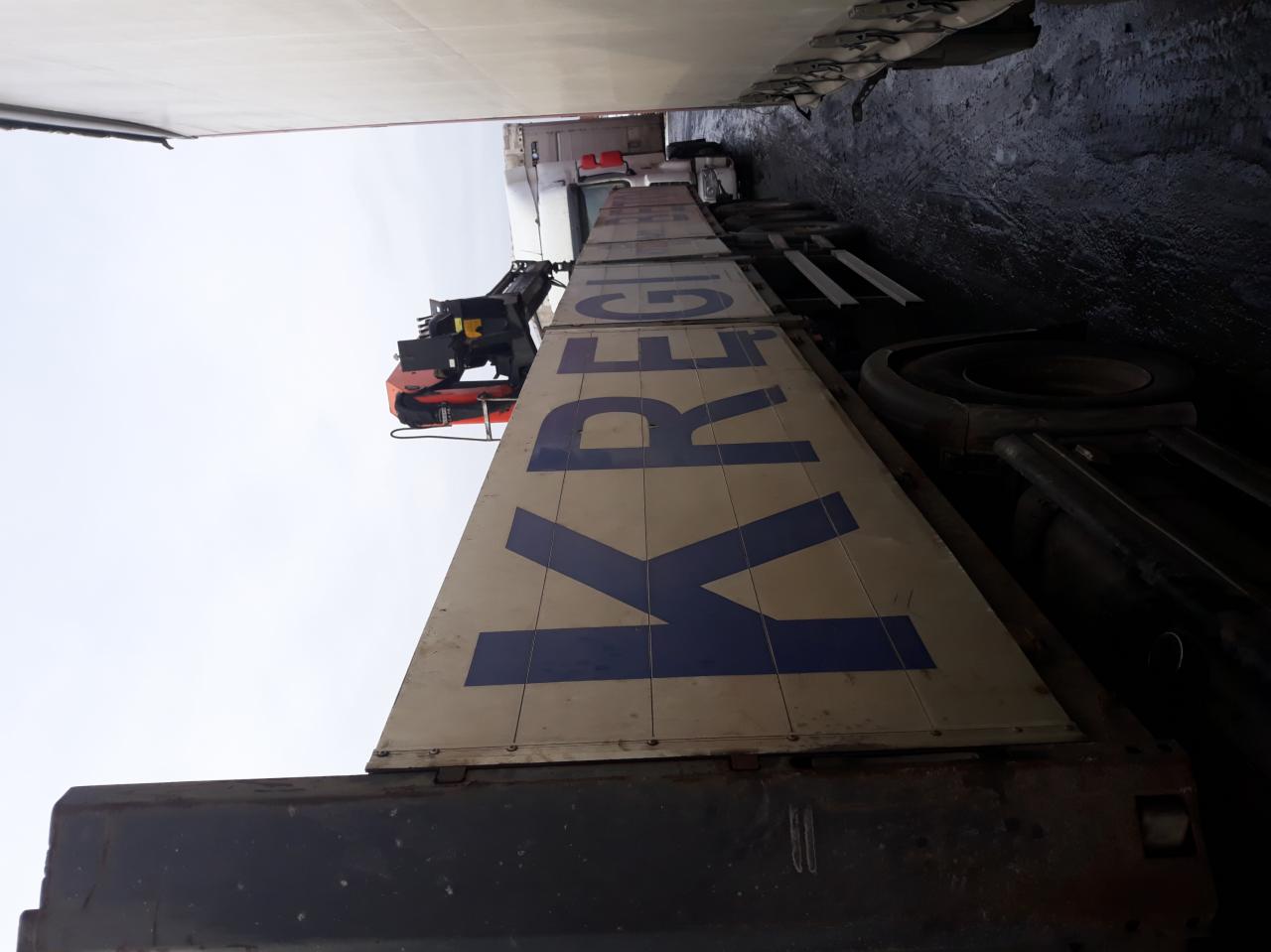 Ciężarówka z pomarańczowym dźwigiem hydraulicznym, widok z dołu na naczepę z napisem KRE, zaparkowana wewnątrz hali.