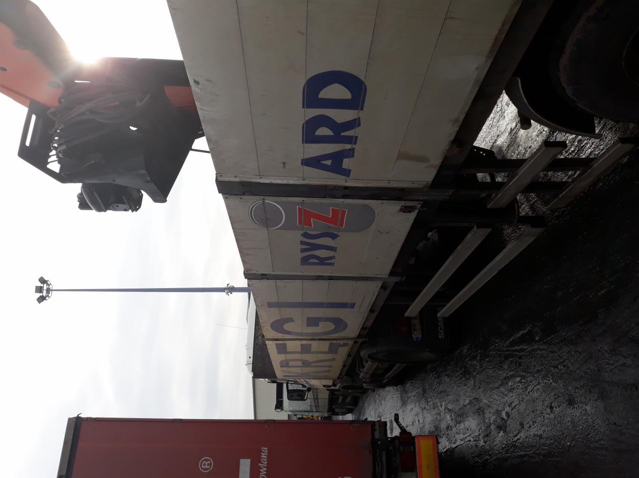 Ciężarówka z dźwigiem na tle lamp oświetleniowych, widoczne logo firmy transportowej.